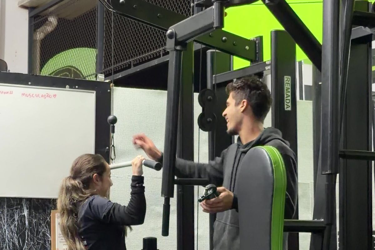 Professor do MyBox corrigindo a postura de um aluno na área de musculação, com aparelhos ao fundo